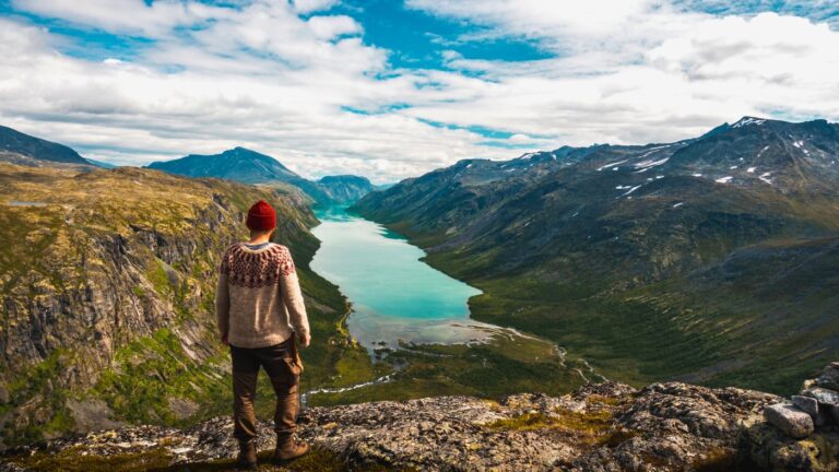 Hiker in red beanie and Nordic sweater admires a scenic view from Gjendetunga peak over Gjende lake in Jotunheimen National Park, Norway.