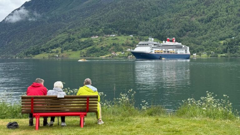 Three people in bright coats on a bench in Skjolden looking at a cruise ship in the fjord. Photo: David Nikel.